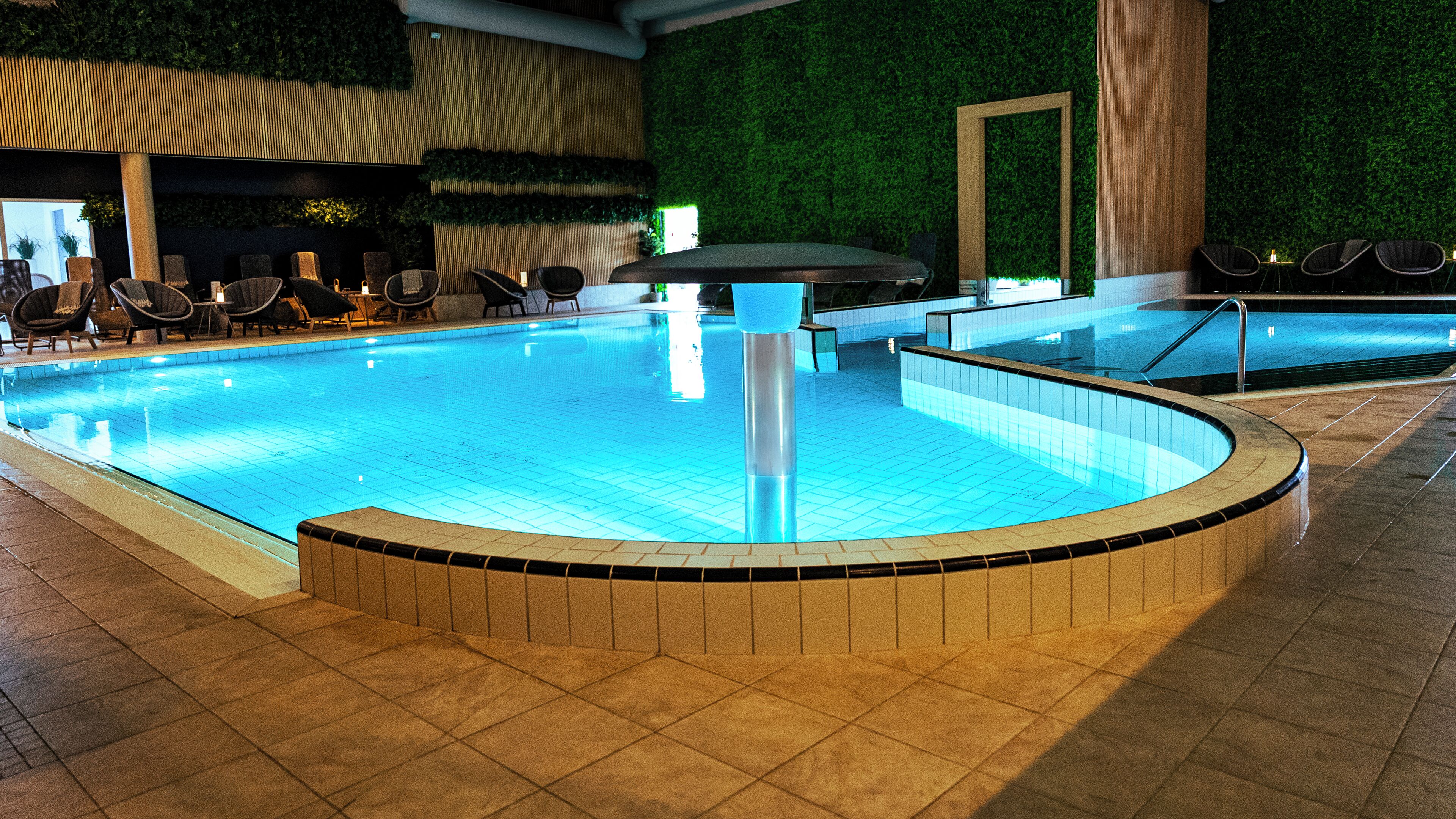 An indoor swimming pool with a tiled edge, surrounded by lounge chairs and a green wall.