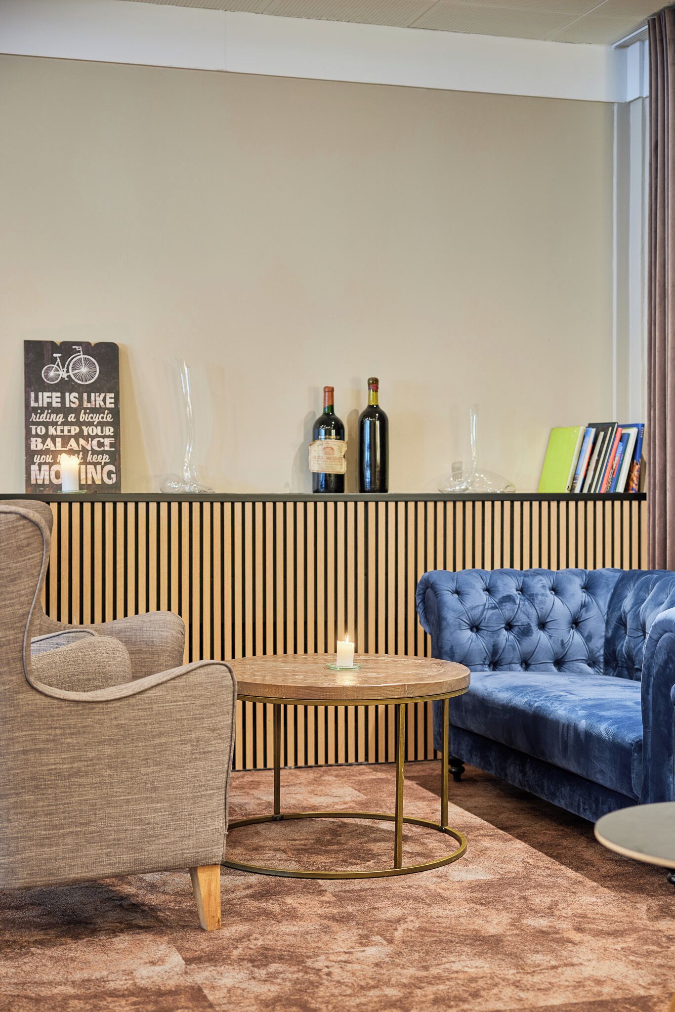 Una sala moderna con un sofá azul, una mesa de centro redonda de madera y un sillón gris. Un separador de pared de listones de madera delimita la zona de estar de una estantería con libros y objetos decorativos.