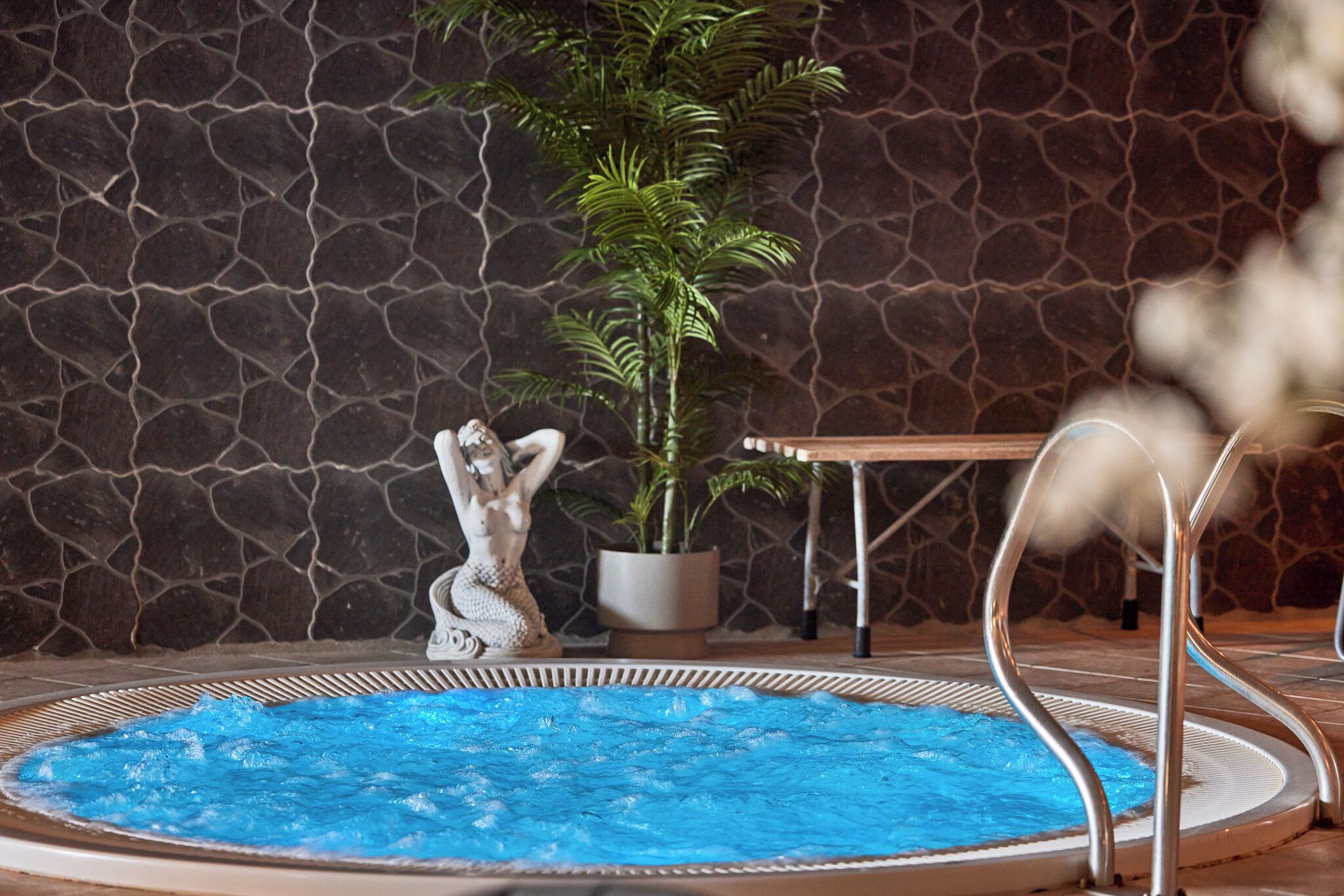 A hot tub with a blue water surface, surrounded by a stone wall and a potted plant.