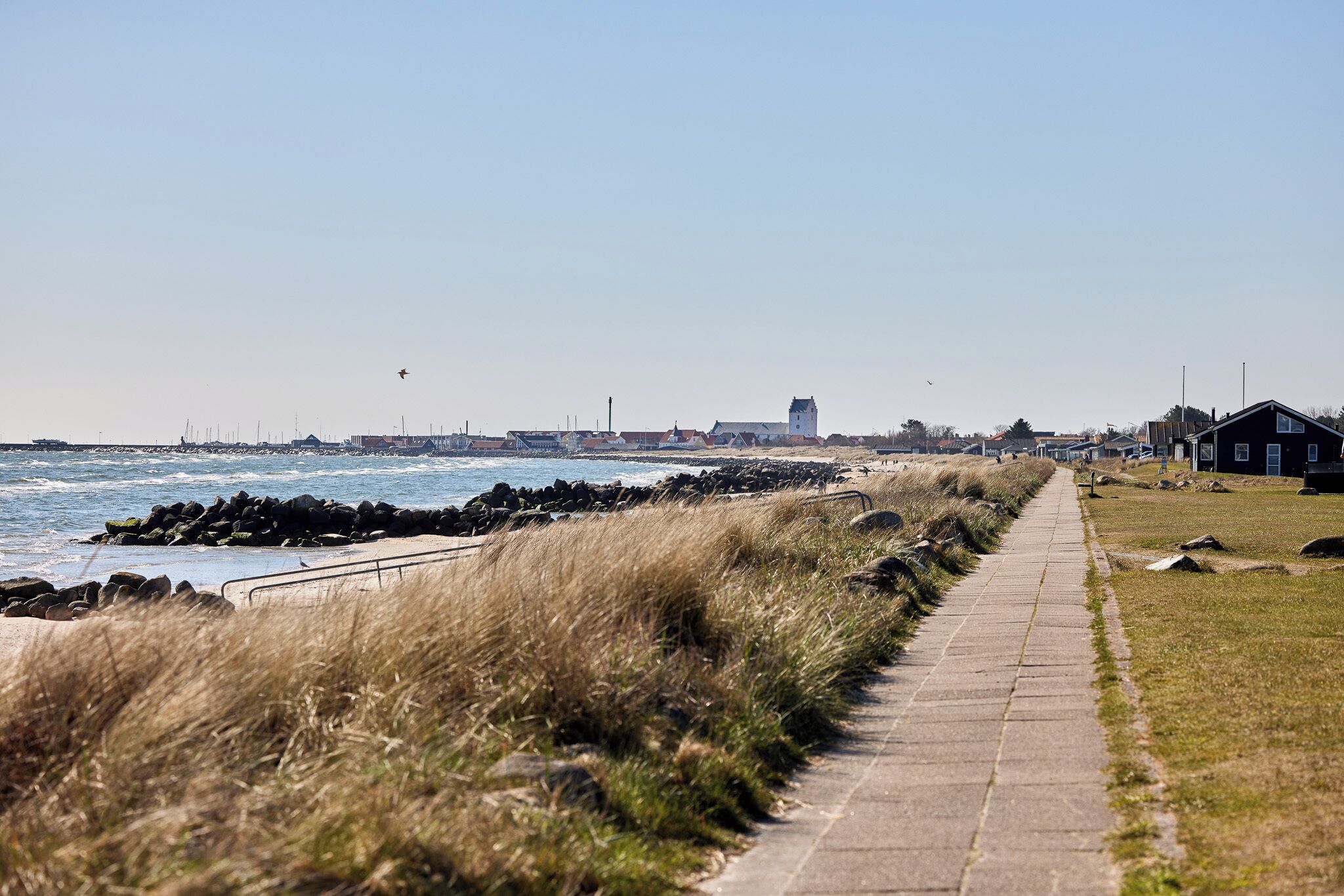 A coastal walkway with a stone path, grassy areas, and a view of the sea and distant buildings.
