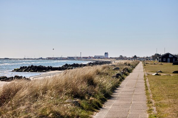 Un paseo marítimo con sendero de piedra, zonas verdes y vistas al mar y edificios a la distancia.