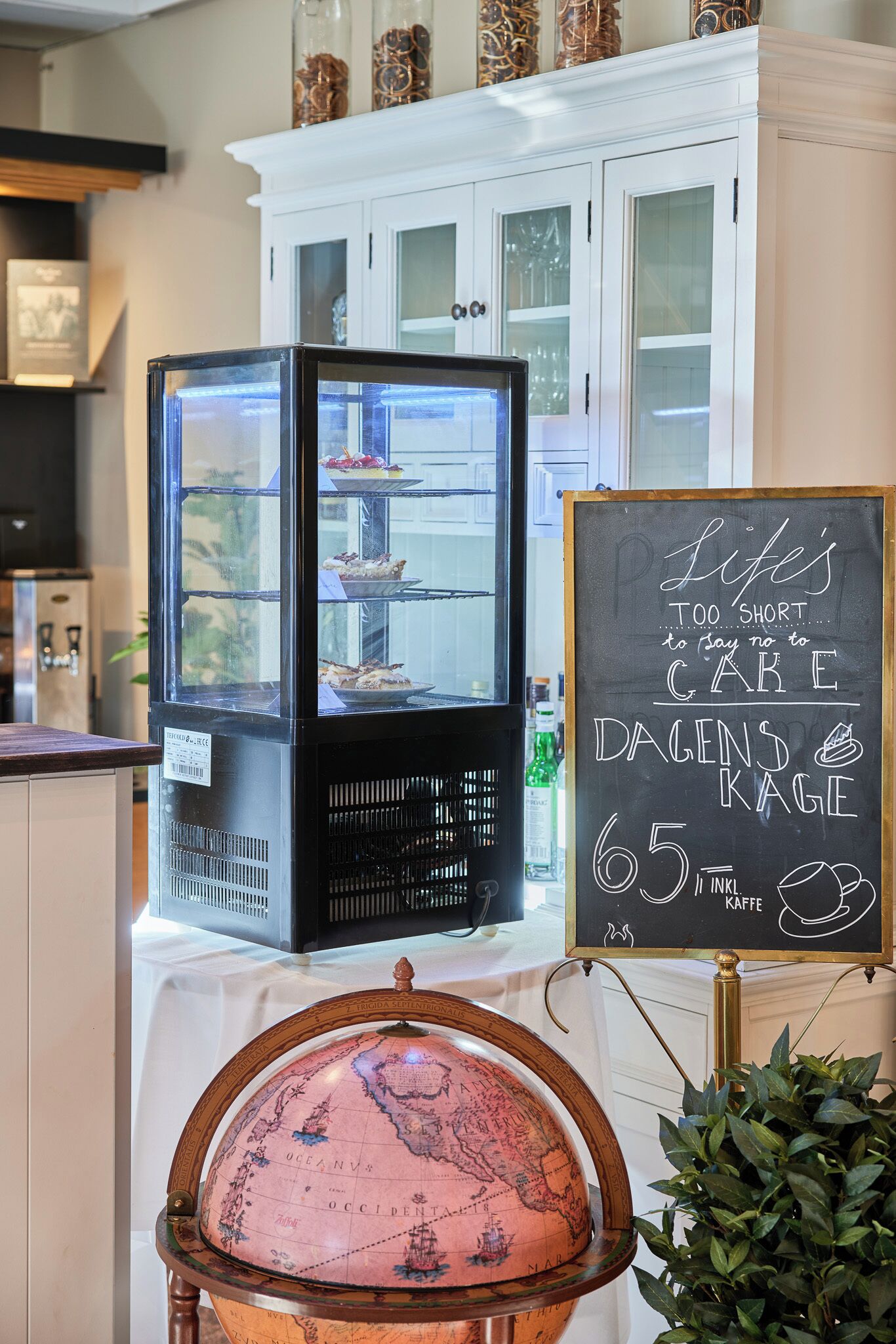 A bakery display with a chalkboard sign advertising cakes and coffee, a globe, and a refrigerator with pastries.