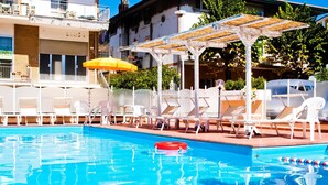Une piscine entourée de bâtiments, avec des chaises longues blanches et un parasol jaune.