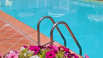 A swimming pool with a tiled deck, surrounded by lounge chairs and umbrellas, and a flower bed with pink flowers in the foreground.