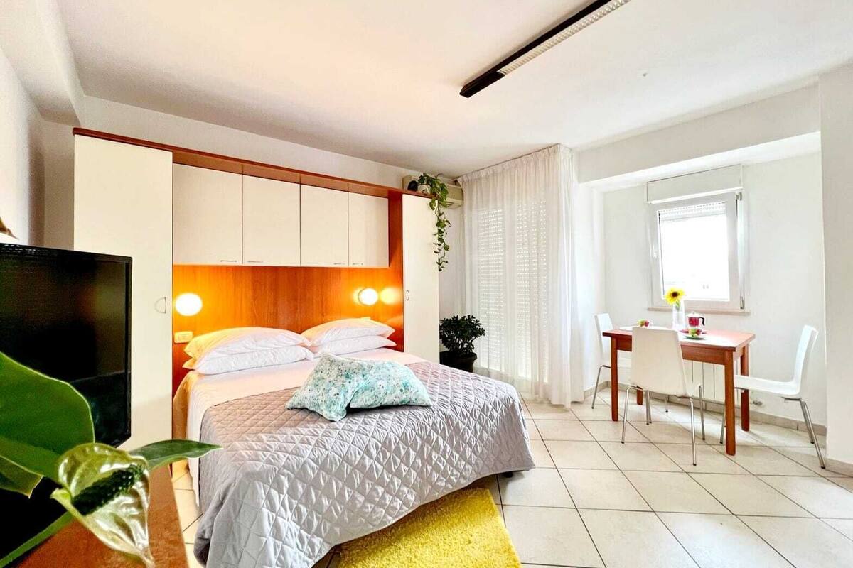 A bedroom with a bed, a desk, a chair, a TV, and a window.