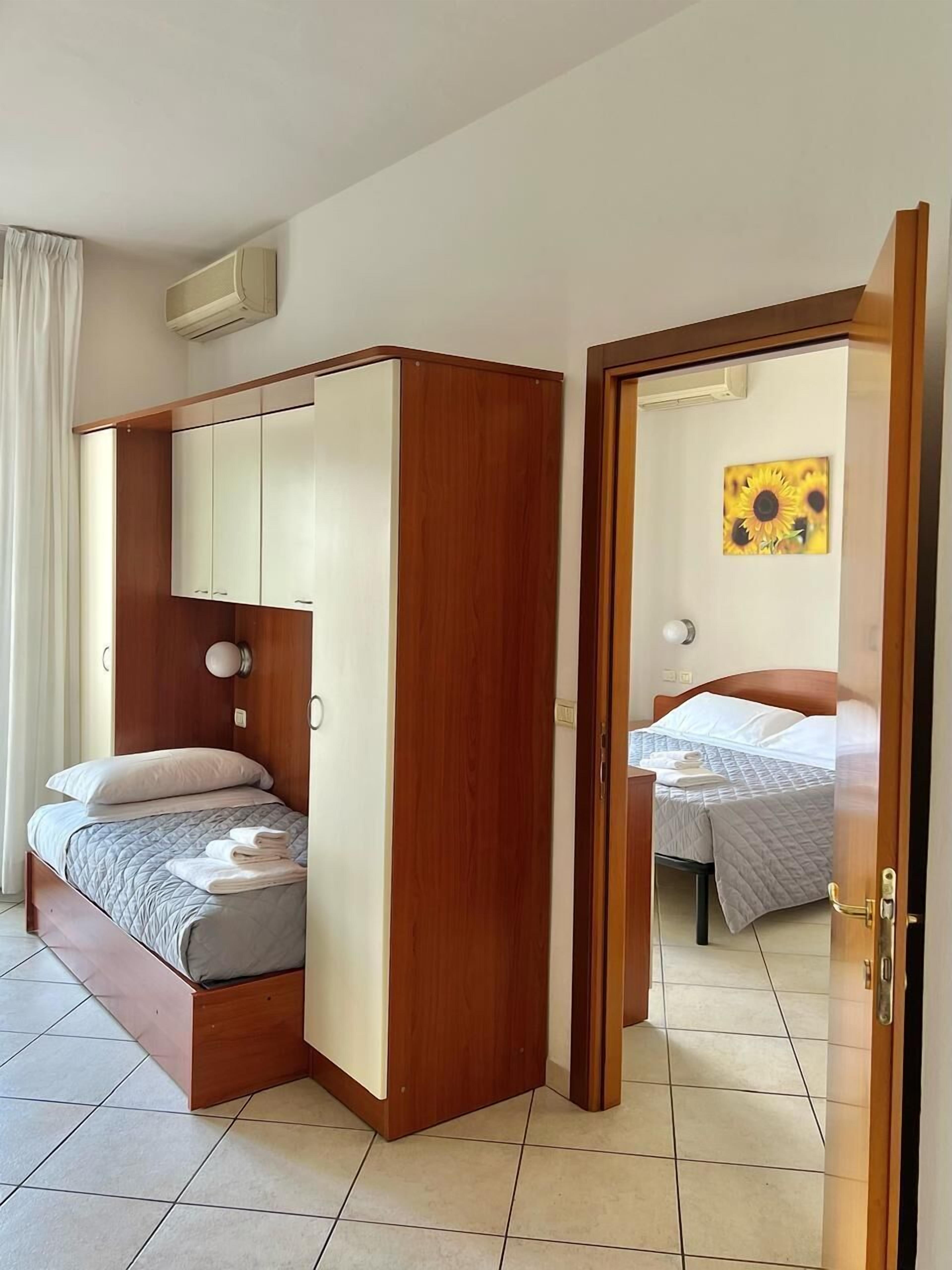 Una habitación con una cama, un armario de madera, una puerta que da a otra habitación y un aire acondicionado instalado en la pared.