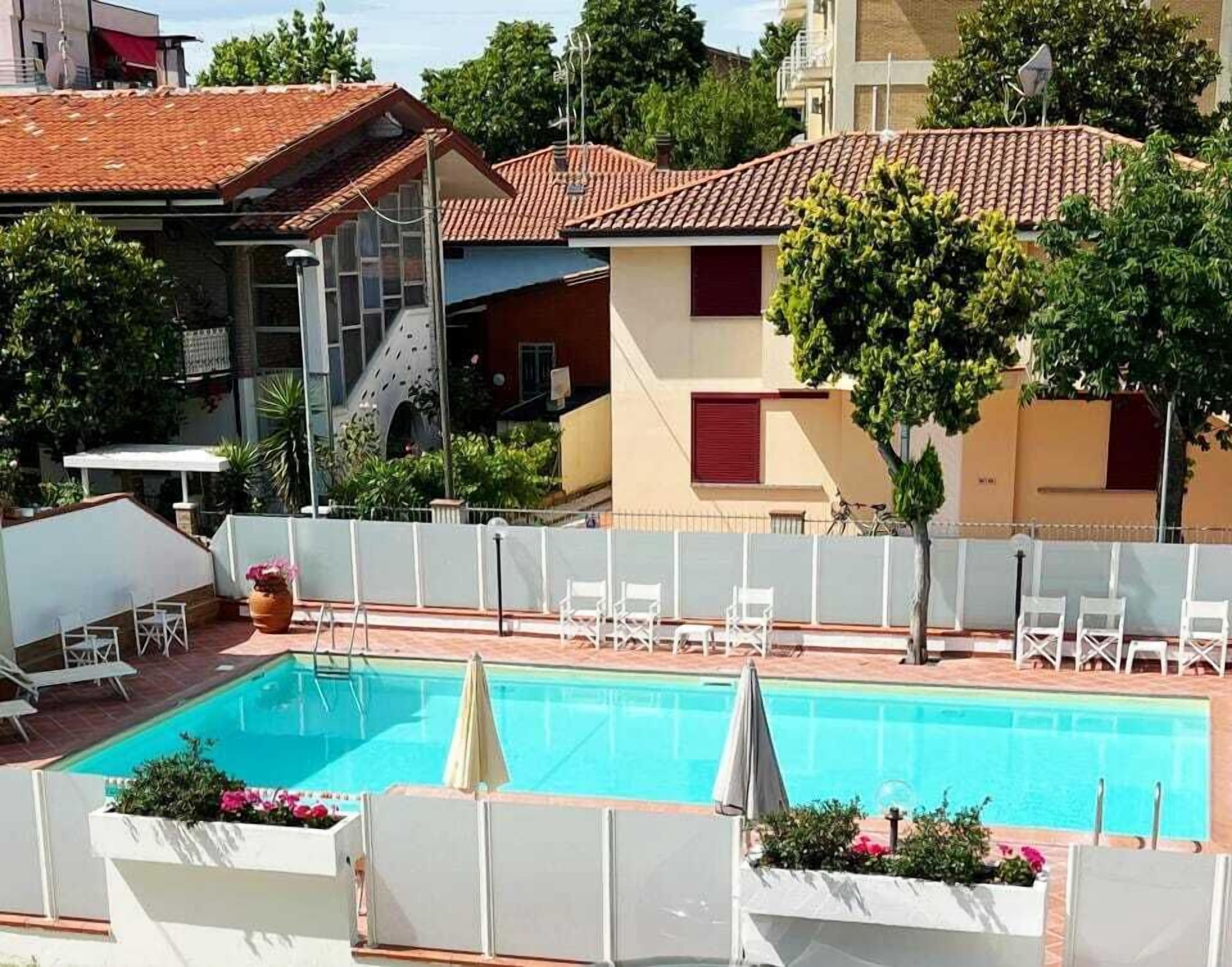 Una piscina con sillas de descanso y sombrillas, rodeada por una valla blanca, con edificios y árboles al fondo.