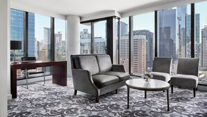 Una habitación de hotel moderna con un ventanal grande, un sofá gris, una mesita redonda y una alfombra estampada.