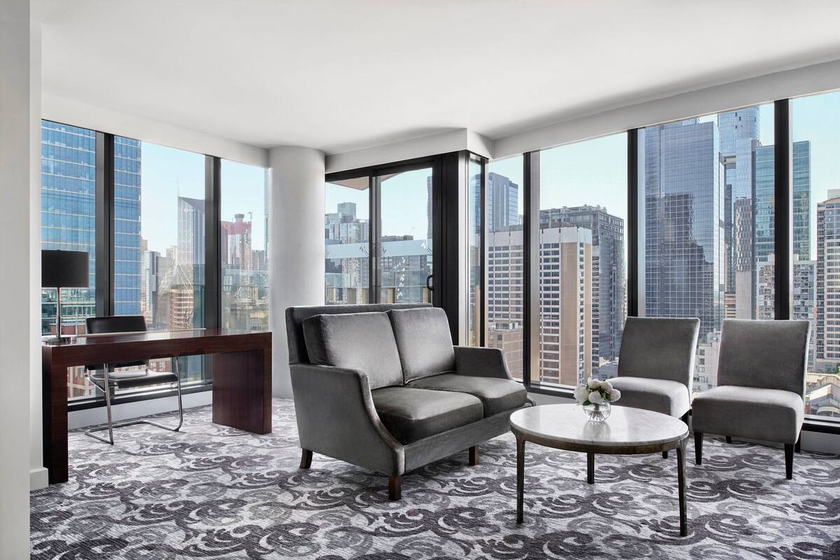 A modern hotel room with a large window, a grey sofa, a small round table, and a patterned carpet.