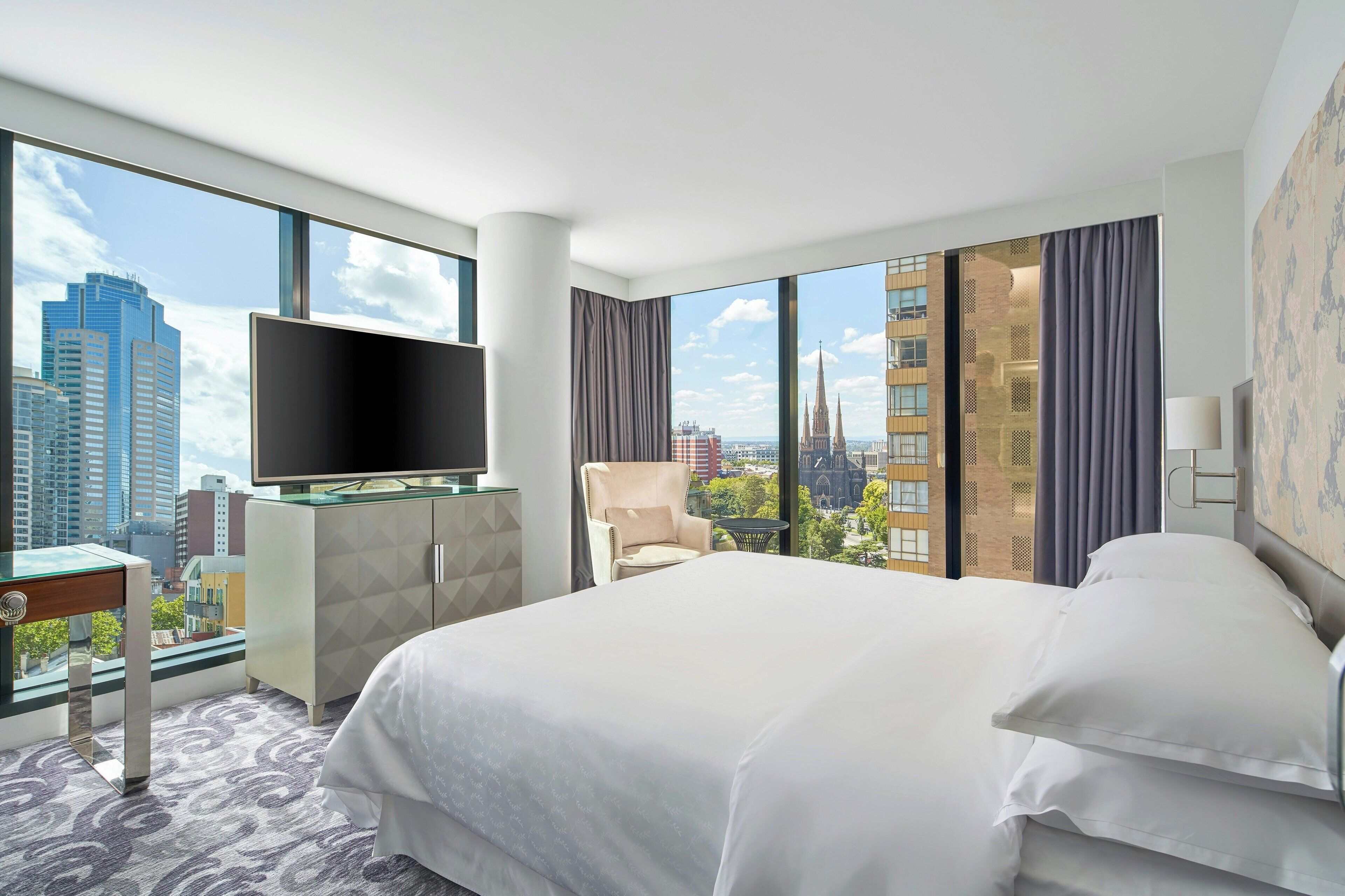 A hotel room with a large bed, a flat-screen TV, a desk, and a view of the cityscape.