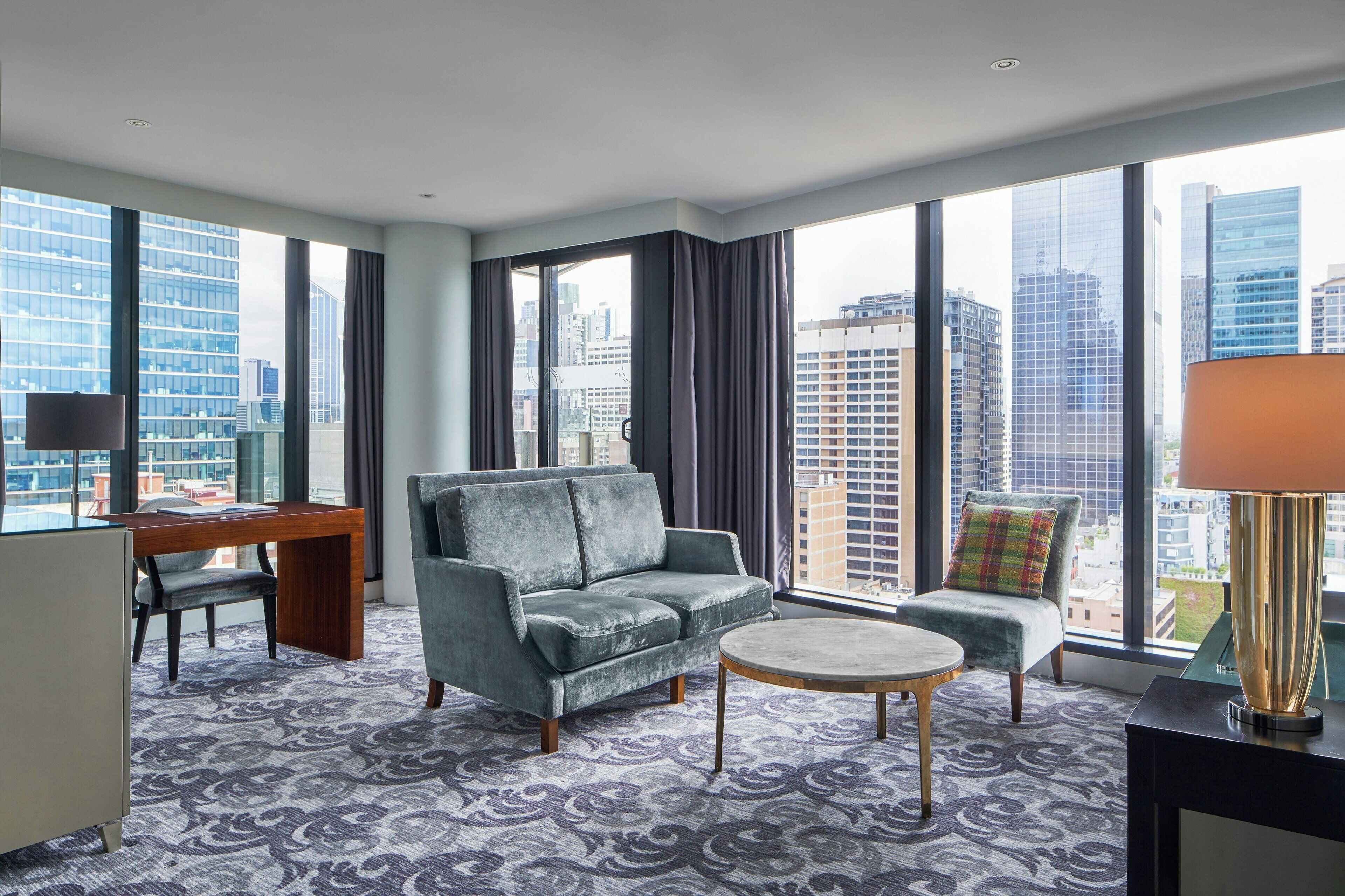Een moderne hotelkamer met een grijze bank, een klein rond tafeltje, een bureau met stoel en uitzicht op de skyline door grote ramen.