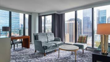 Een moderne hotelkamer met een grijze bank, een klein rond tafeltje, een bureau met stoel en uitzicht op de skyline door grote ramen.