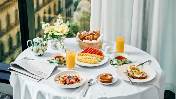 Una mesa de desayuno con croissants, fruta y bebidas, frente a una ventana con vista a la ciudad.