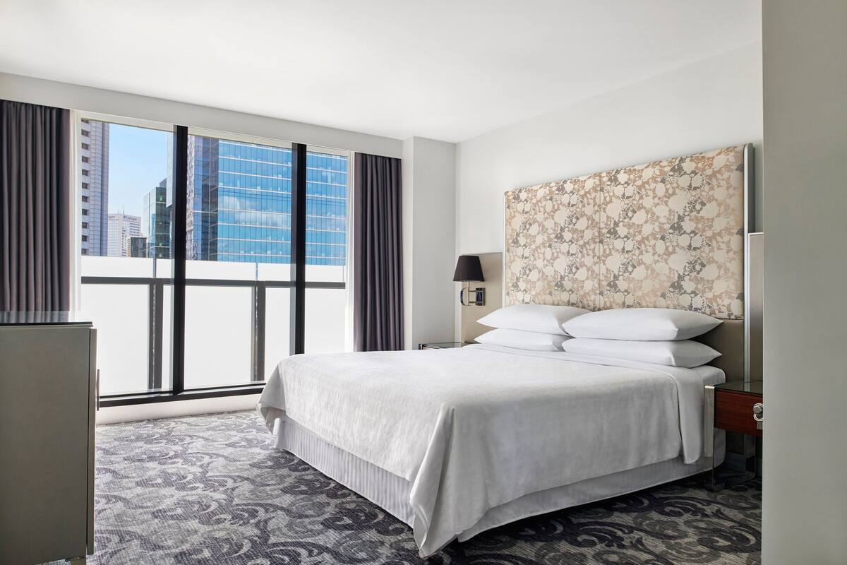 A hotel room with a large bed, a nightstand, a lamp, and a view of the city through large windows.