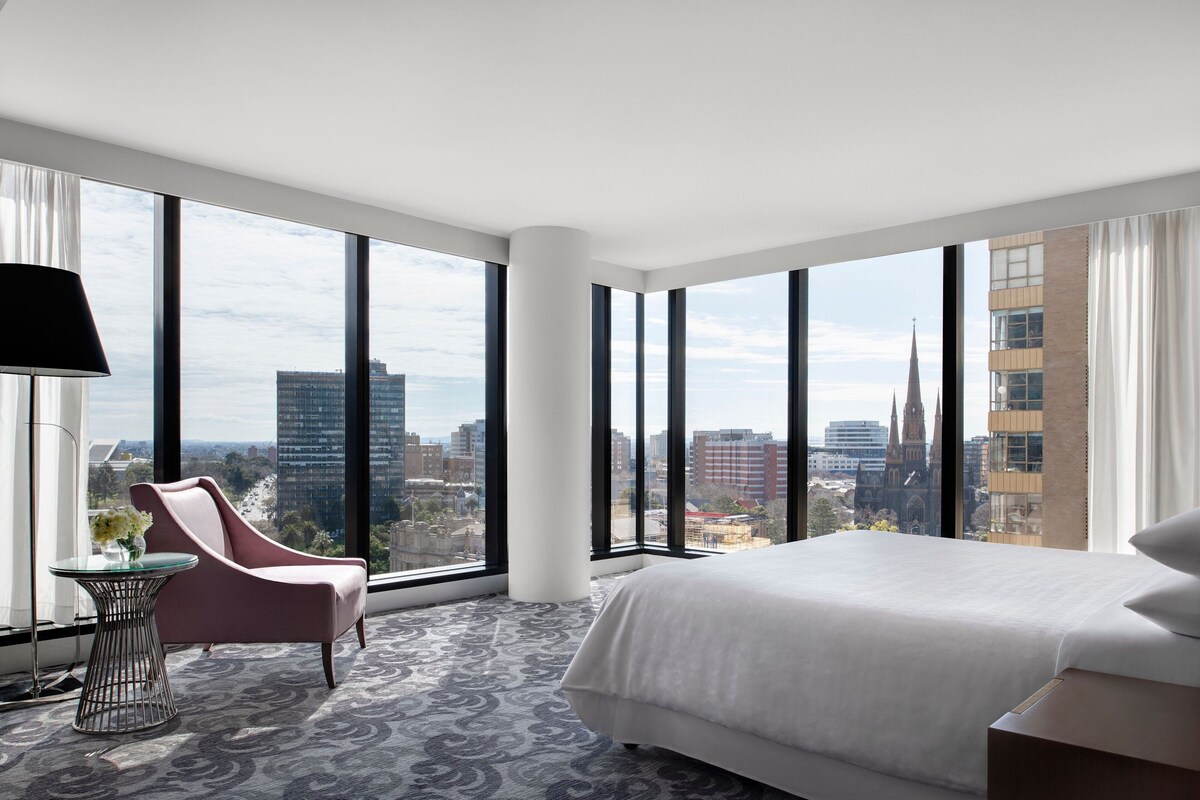 A modern hotel room with a large bed, a pink armchair, a small table with a vase, and large windows offering a city view.