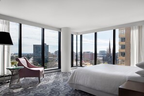 A modern hotel room with a large bed, a pink armchair, a small table with a vase, and large windows offering a city view. - Sheraton Melbourne Hotel (Melbourne)