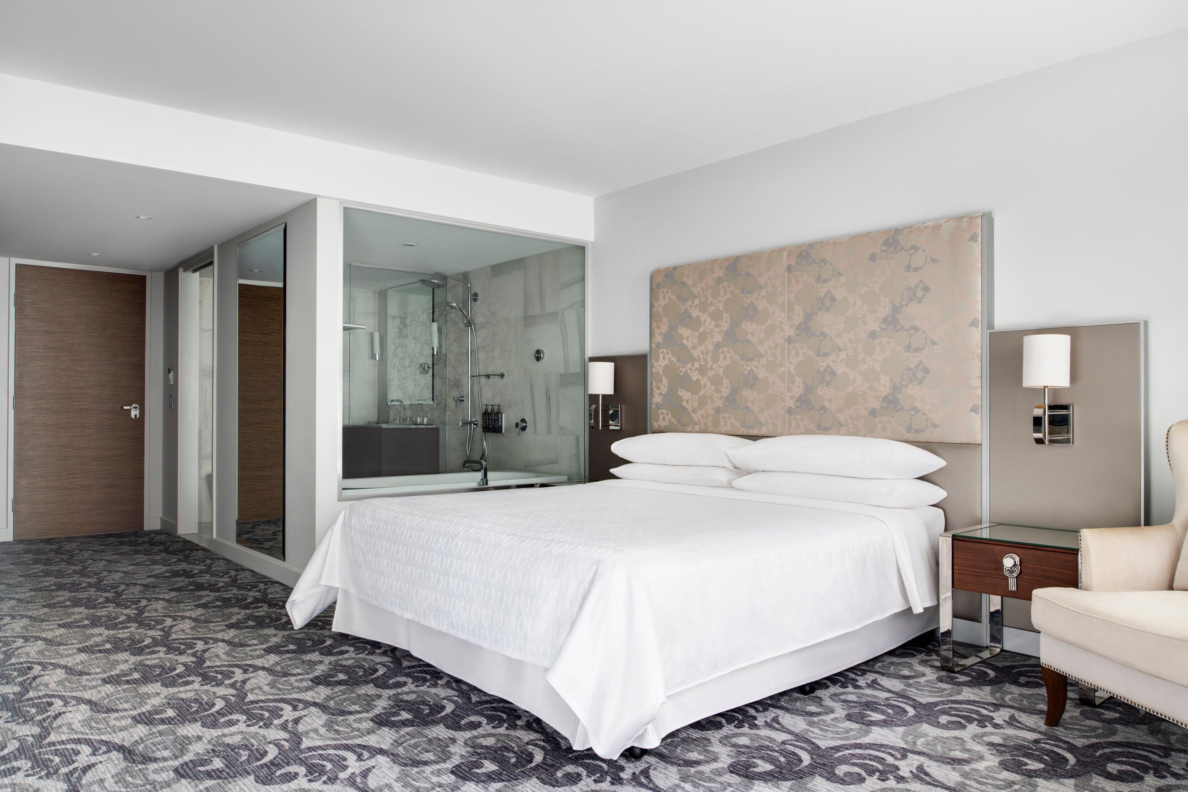 A modern hotel room with a large bed, a glass shower enclosure, a bedside table with a lamp, and a patterned carpet.
