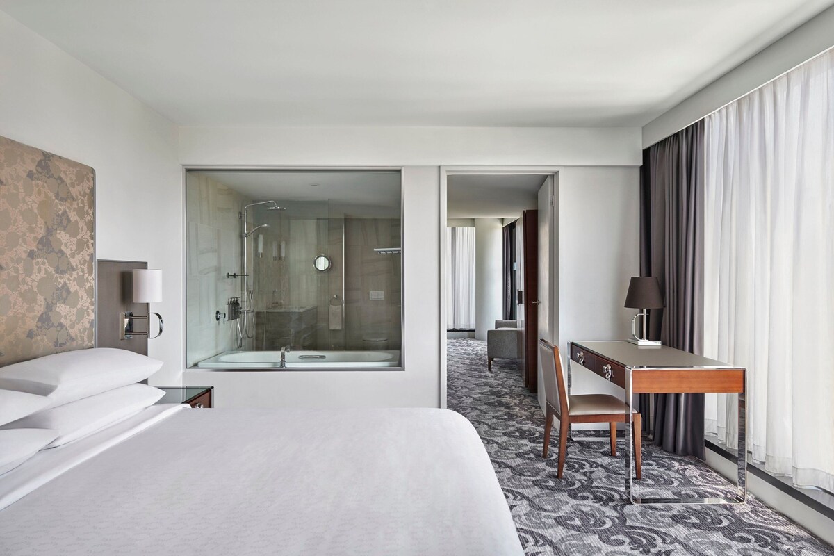 A hotel room with a large bed, a desk, a chair, a desk lamp, a glass shower enclosure, and a patterned carpet.