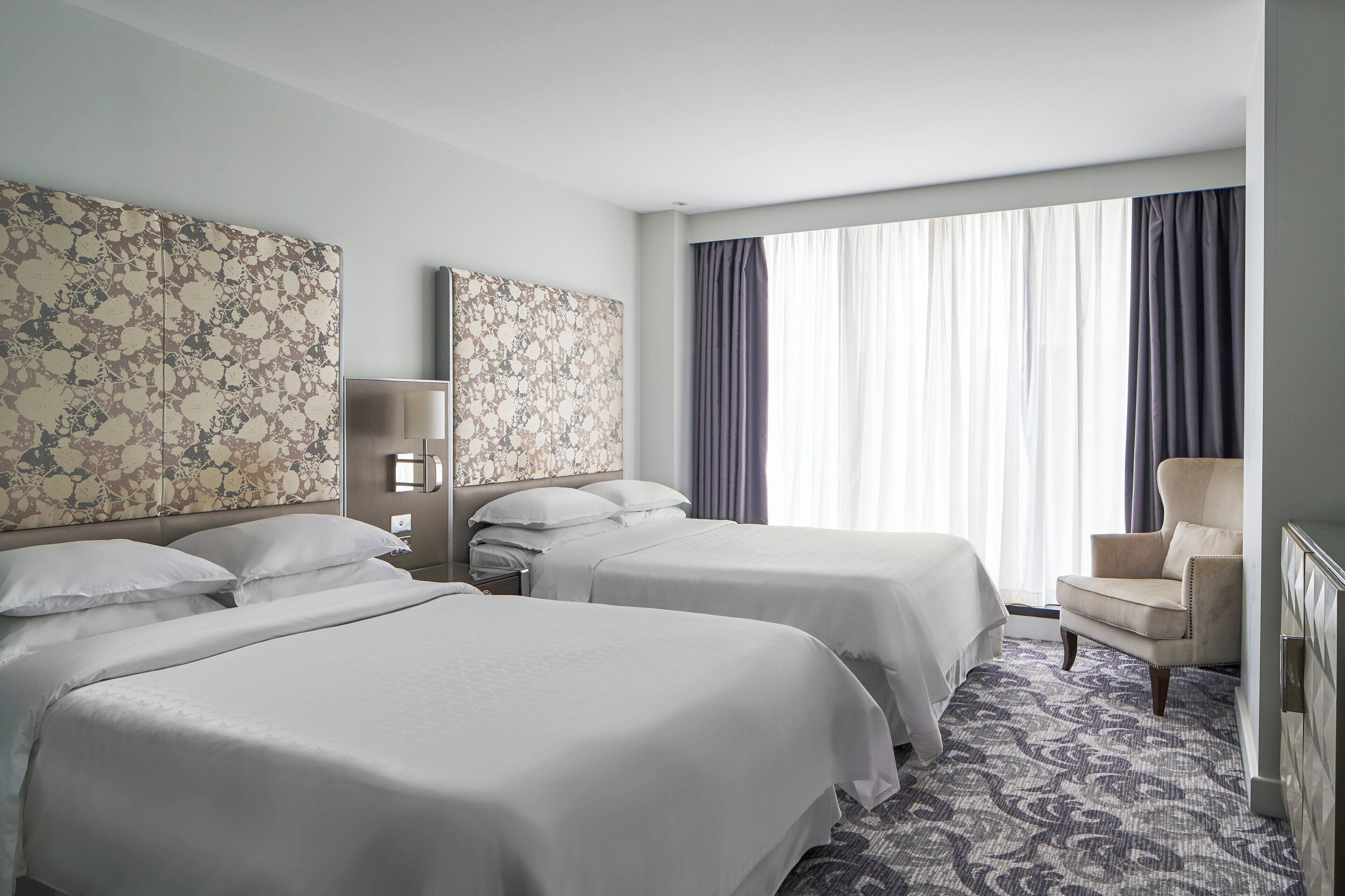 A hotel room with two beds, a chair, a large window with curtains, and a patterned carpet.