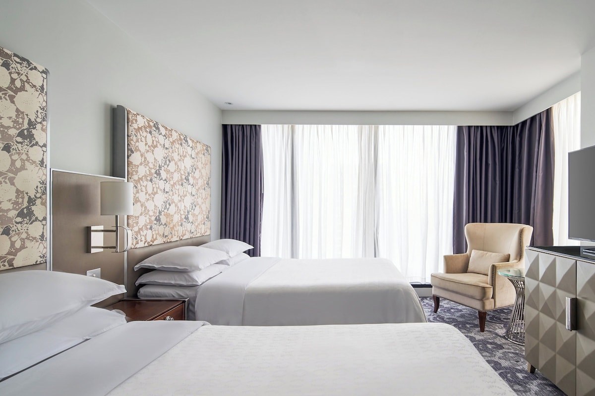 A hotel room with two beds, a chair, a TV, and a large window with curtains.