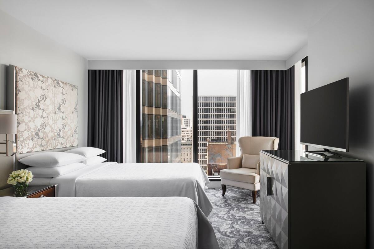 A hotel room with two beds, a TV, a desk, and a view of the cityscape.
