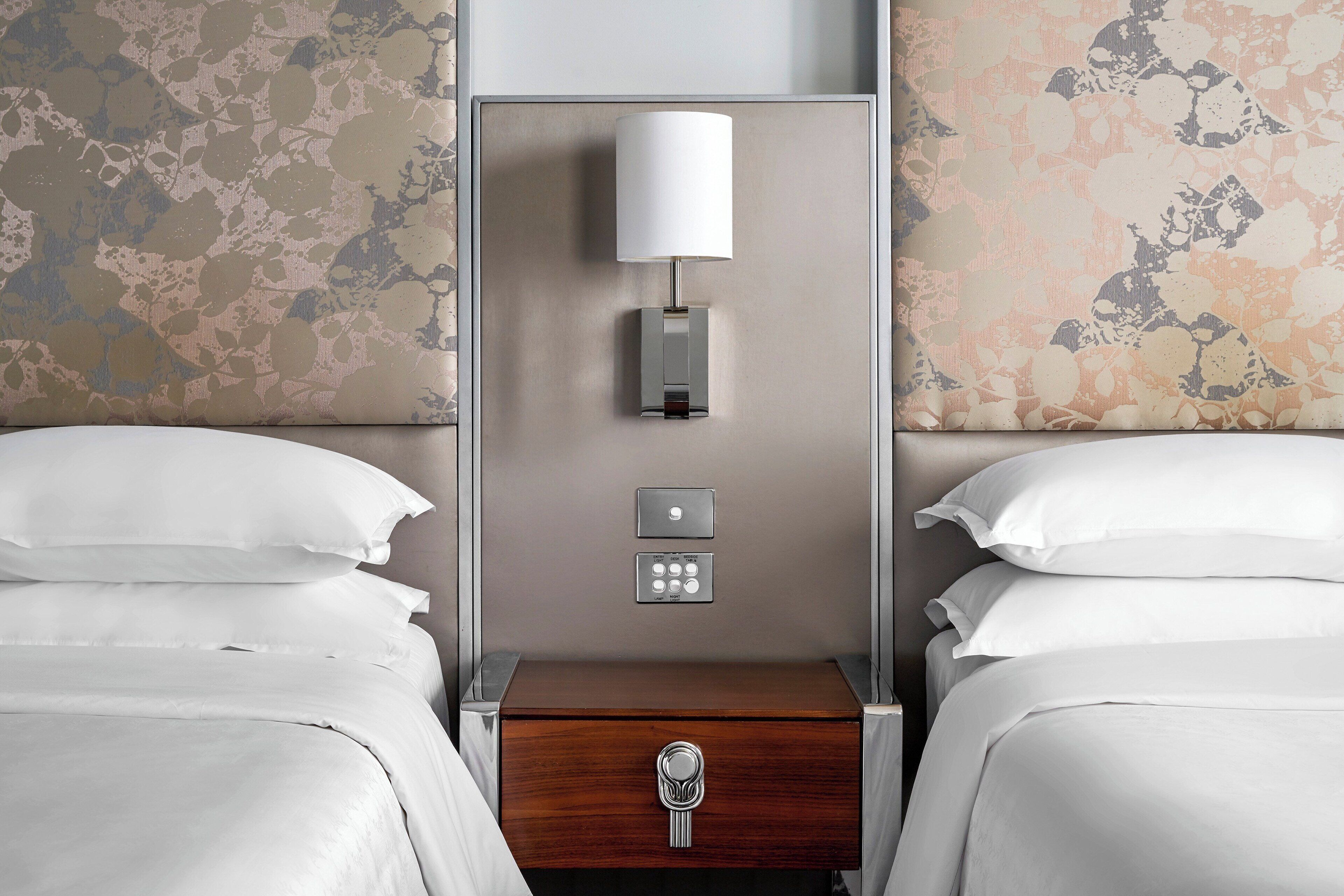 A hotel room with two beds, a bedside table with a lamp, a control panel, and a wooden drawer.