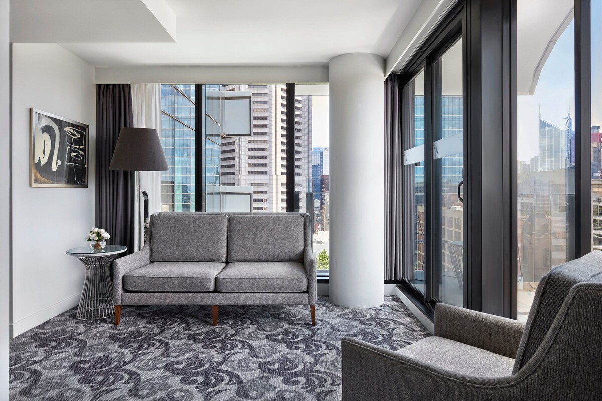 A modern hotel room with a grey sofa, a small side table with a vase, and a large window offering a city view.
