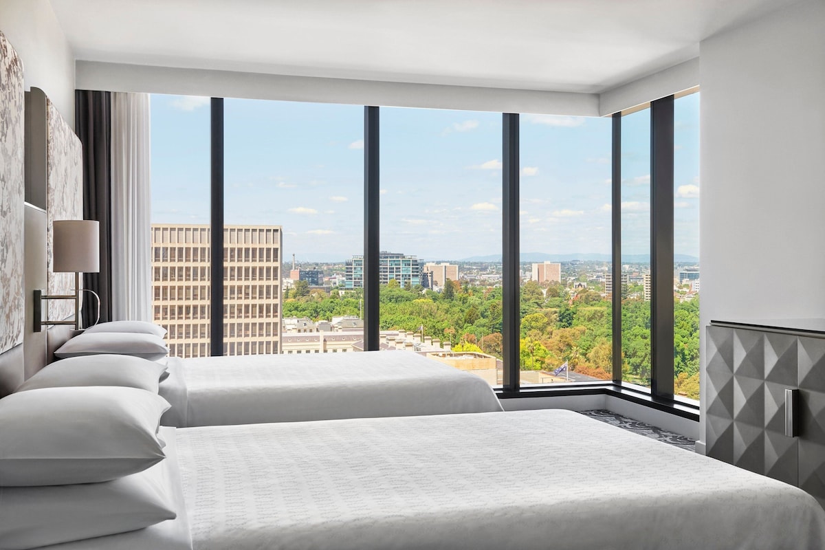 A hotel room with two beds, large windows, and a city view.