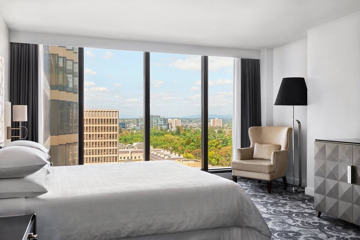 A hotel room with a large bed, a chair, a desk, and a view of the cityscape through large windows.