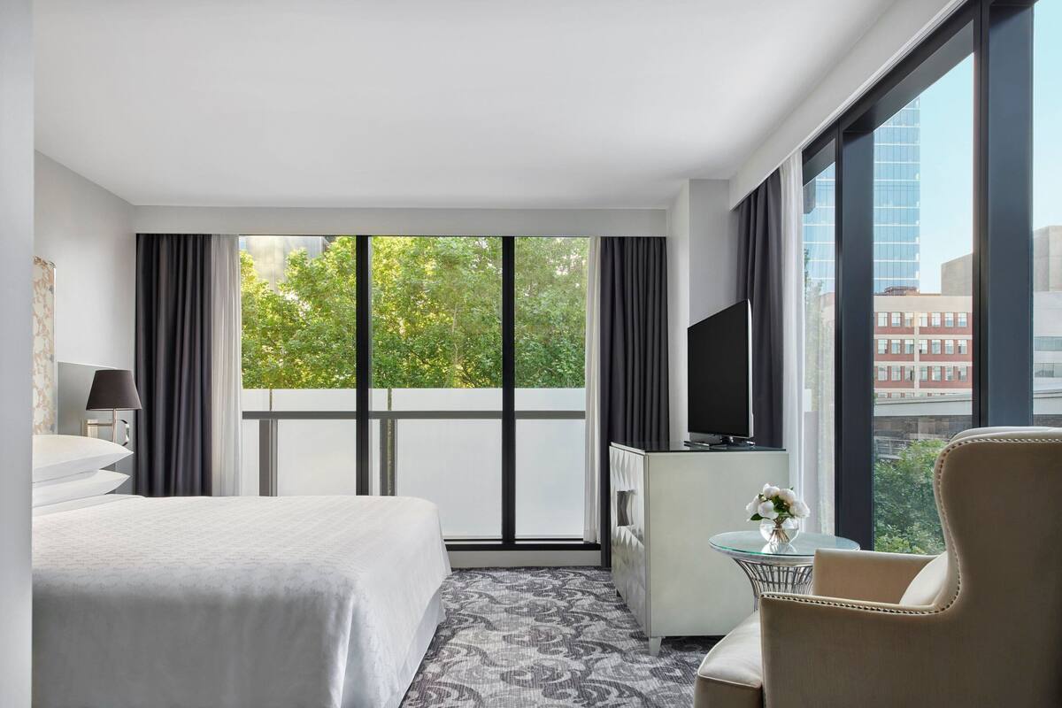 A hotel room with a large bed, a TV, a small table with a vase, and a chair.