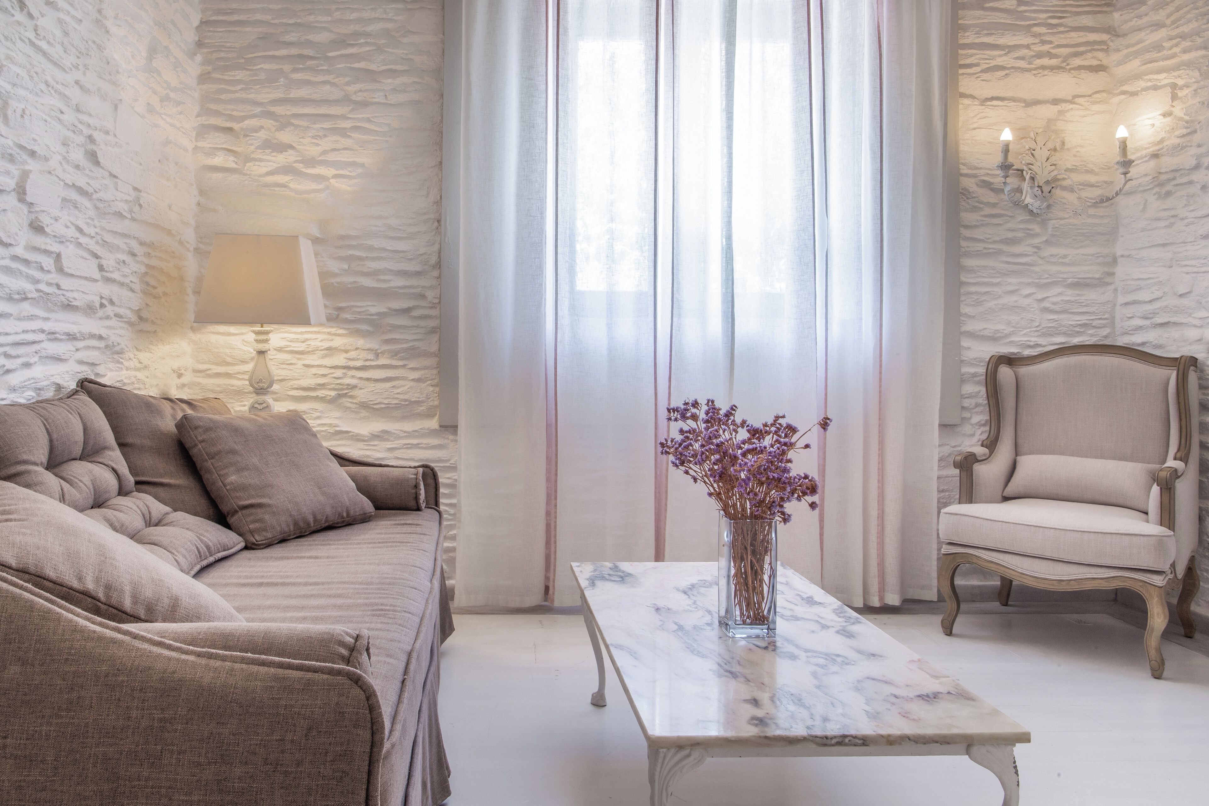 A room with a stone wall, a sofa, a chair, a table with a vase of flowers, and a lamp.