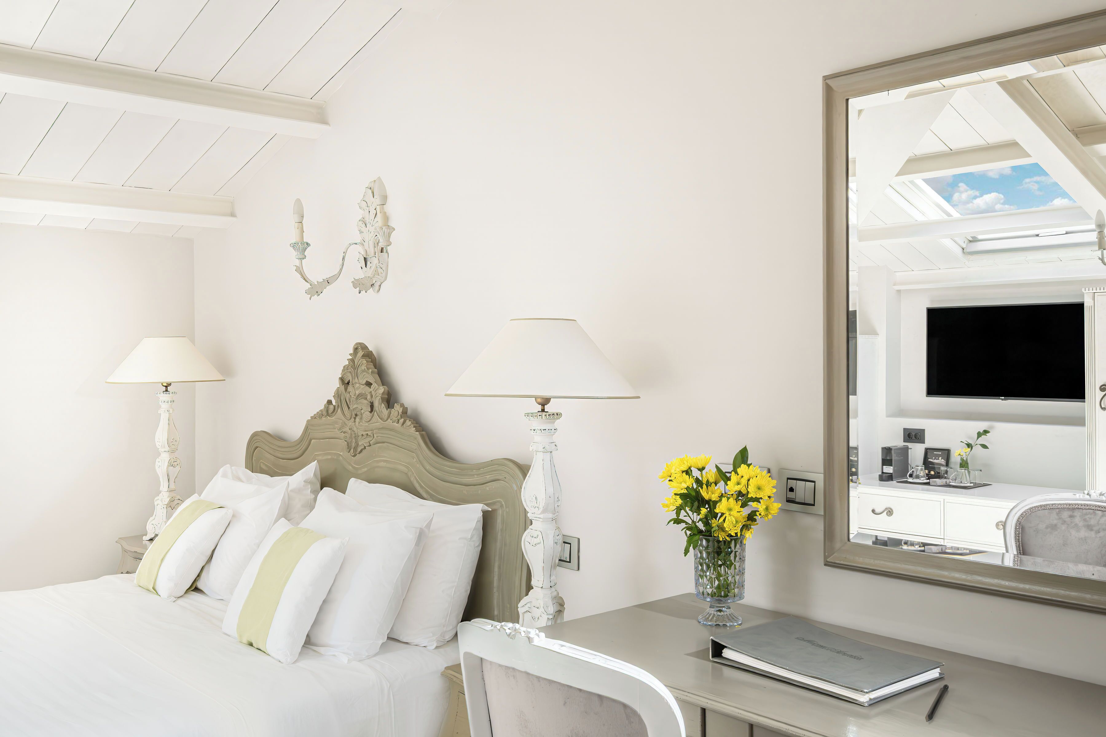 Hotelkamer met een bed, twee bedlampen, een bureau met stoel, een vaas met bloemen, een grote spiegel en een televisie.