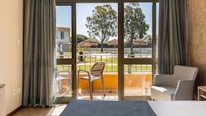 Quarto de hotel com uma janela grande, uma cama, uma cadeira e vista para área externa com guarda-sóis e piscina.
