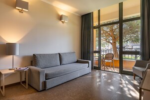 Una habitación de hotel moderna con un sofá gris, una mesita con lámpara y vistas al exterior a través de amplios ventanales.