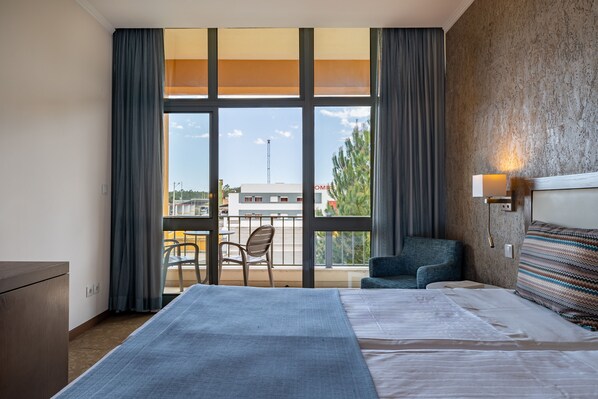 Habitación de hotel con una cama grande, un escritorio, una silla y un balcón con vistas a la ciudad.