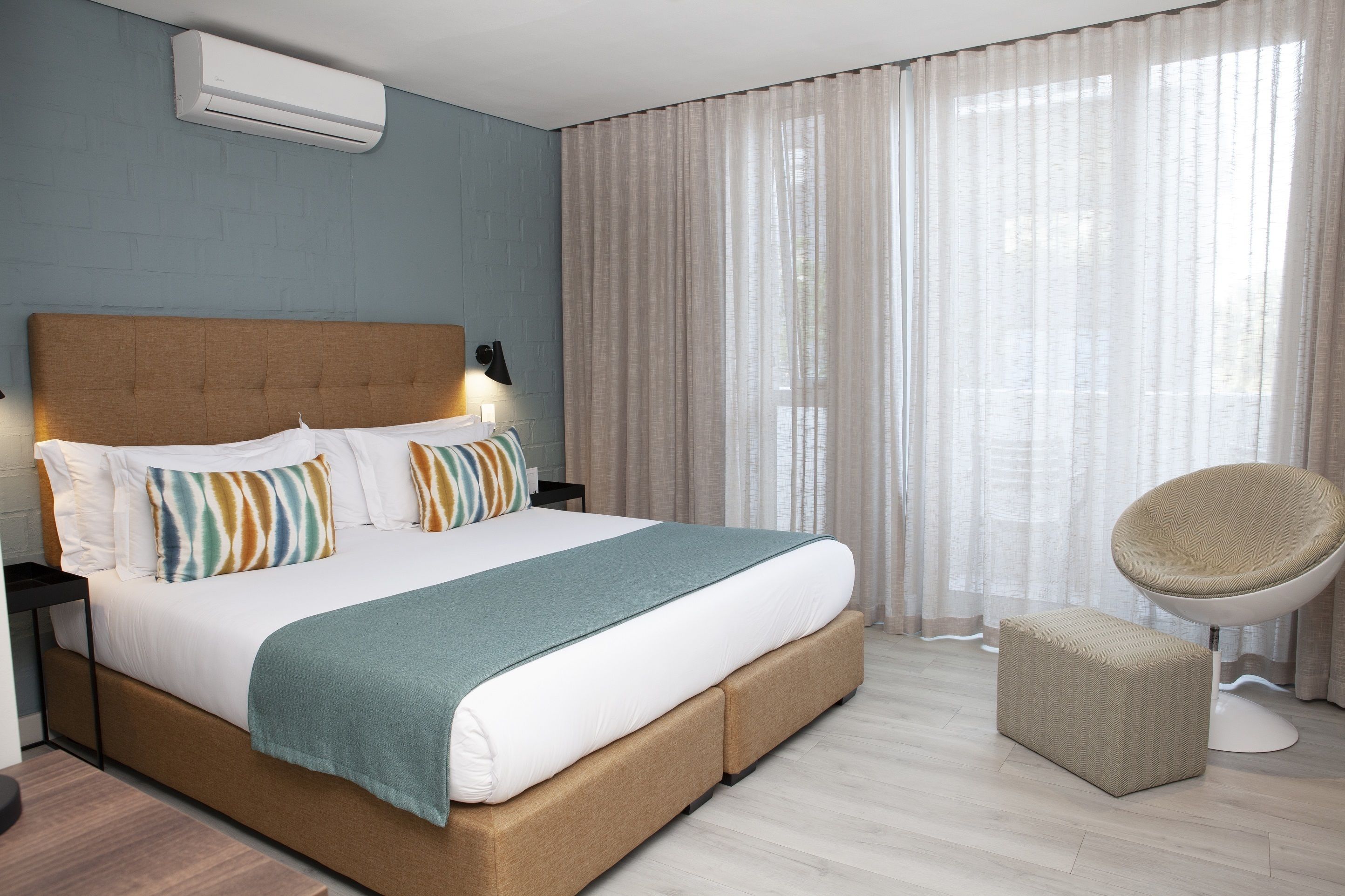 A modern bedroom with a large bed, a bedside table, a chair, and a window with sheer curtains.