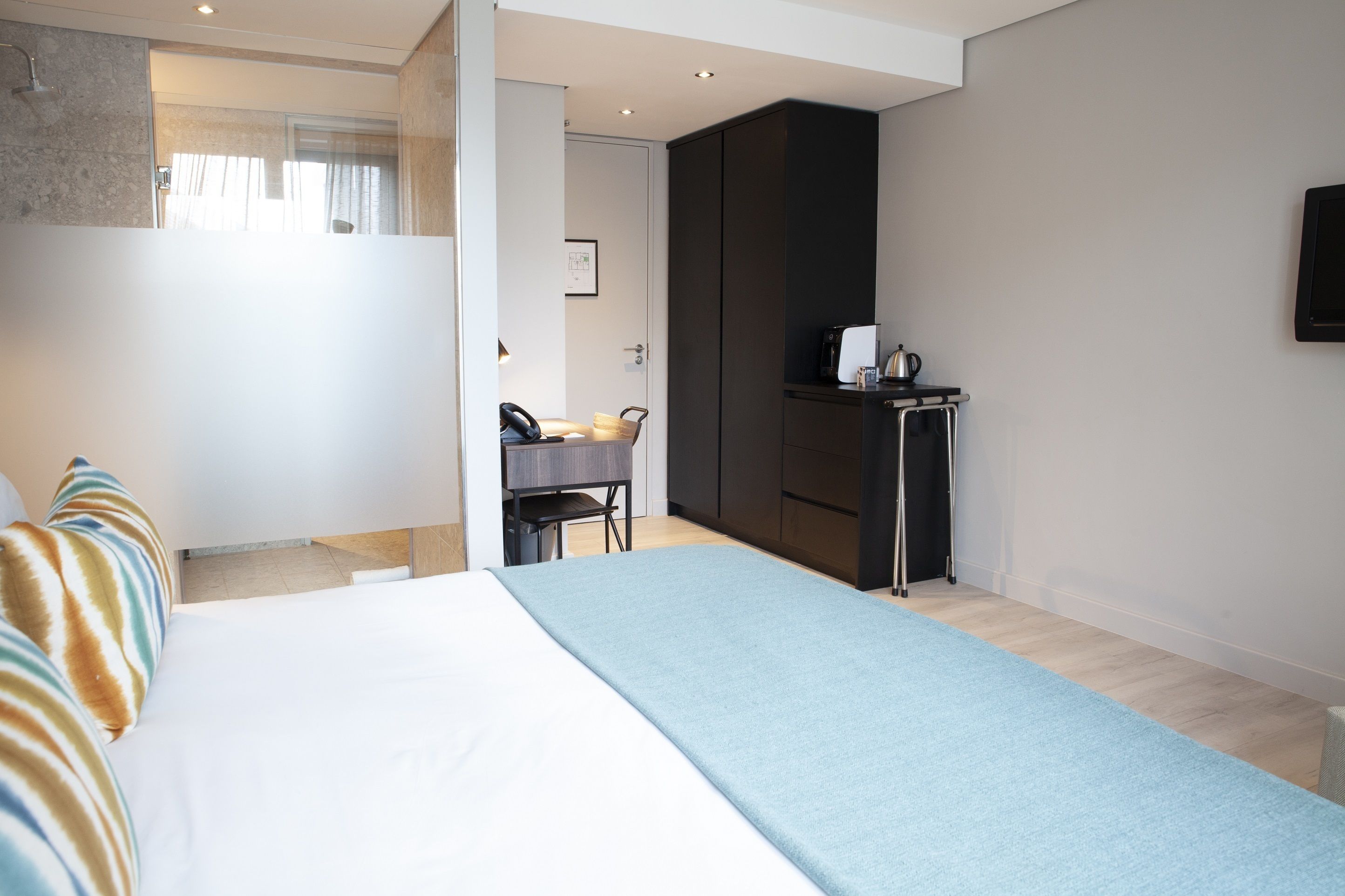 A modern hotel room with a large bed, a desk with a chair, a kitchenette, and a TV mounted on the wall.