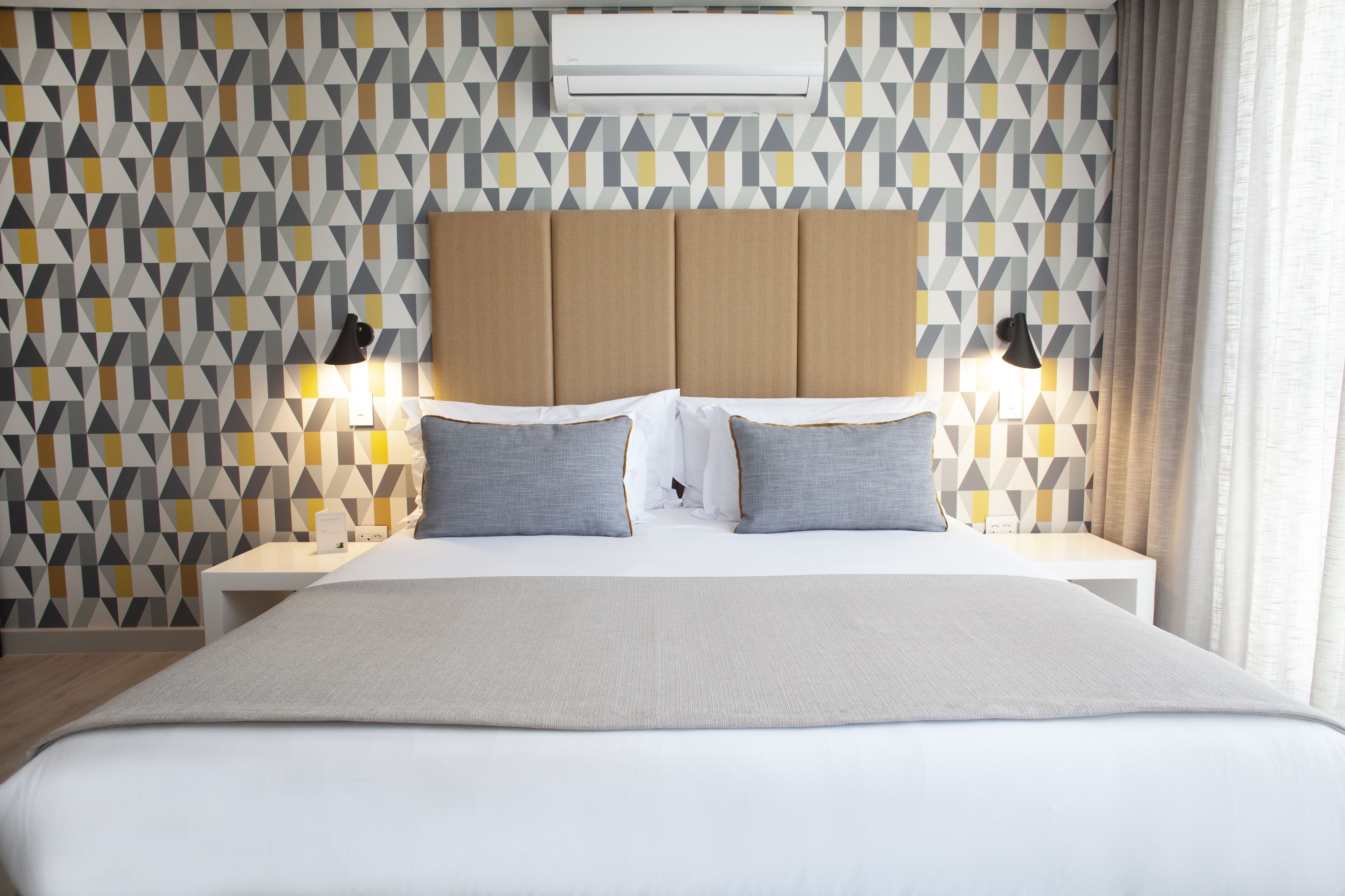 A hotel room with a geometric patterned wall, a neutral-colored headboard, two bedside tables with lamps, and a large bed with a white comforter and gray pillows.