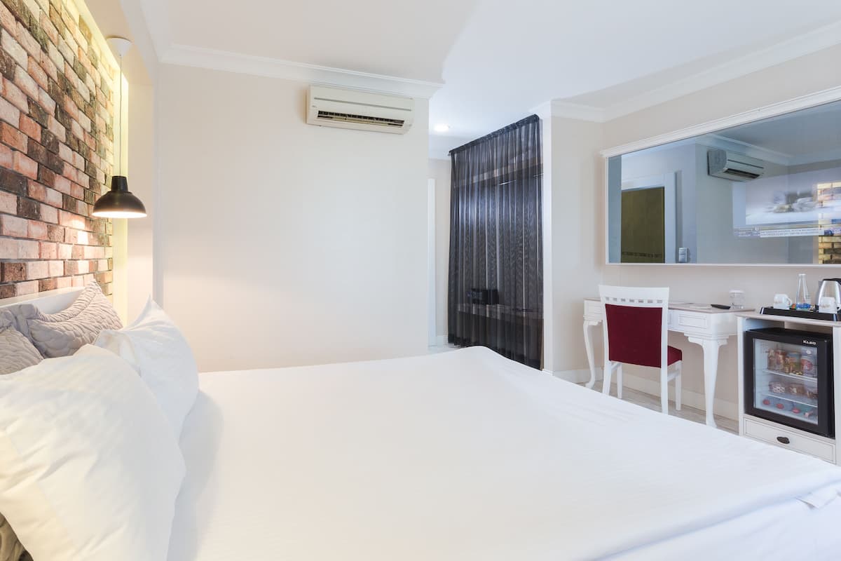 A hotel room with a brick wall, a bed with white bedding, a desk with a chair, and a minibar.
