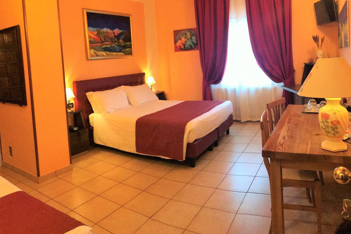 A hotel room with a bed, a desk, a chair, a lamp, and two paintings on the wall.