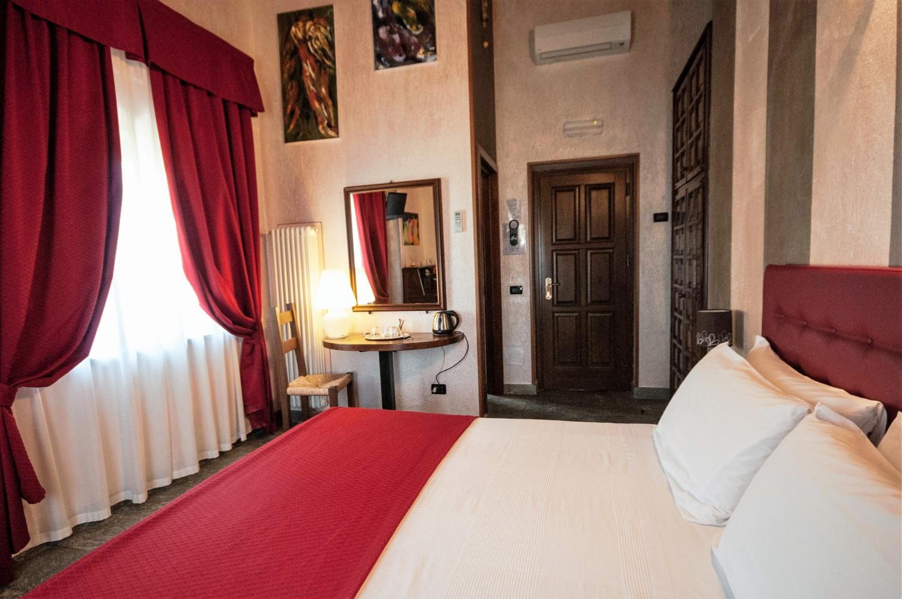 A hotel room with a large bed, red curtains, a small table with a lamp, a mirror, and a door.