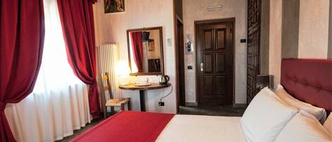 A hotel room with a large bed, red curtains, a small table with a lamp, a mirror, and a door.