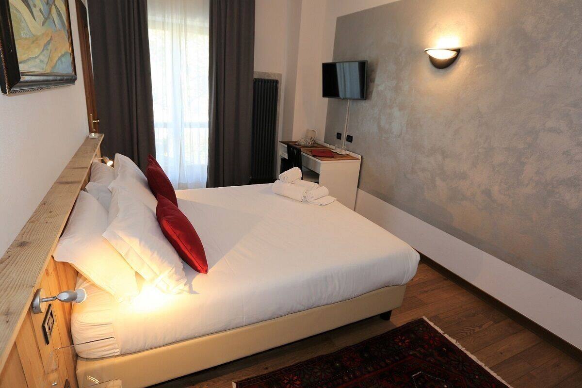 A neatly made bed with white linens and a red pillow, a bedside table with a lamp, a flat-screen TV mounted on the wall, and a wooden floor.