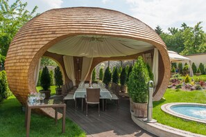 A covered outdoor dining area with a wooden dome structure, surrounded by potted plants and a swimming pool.