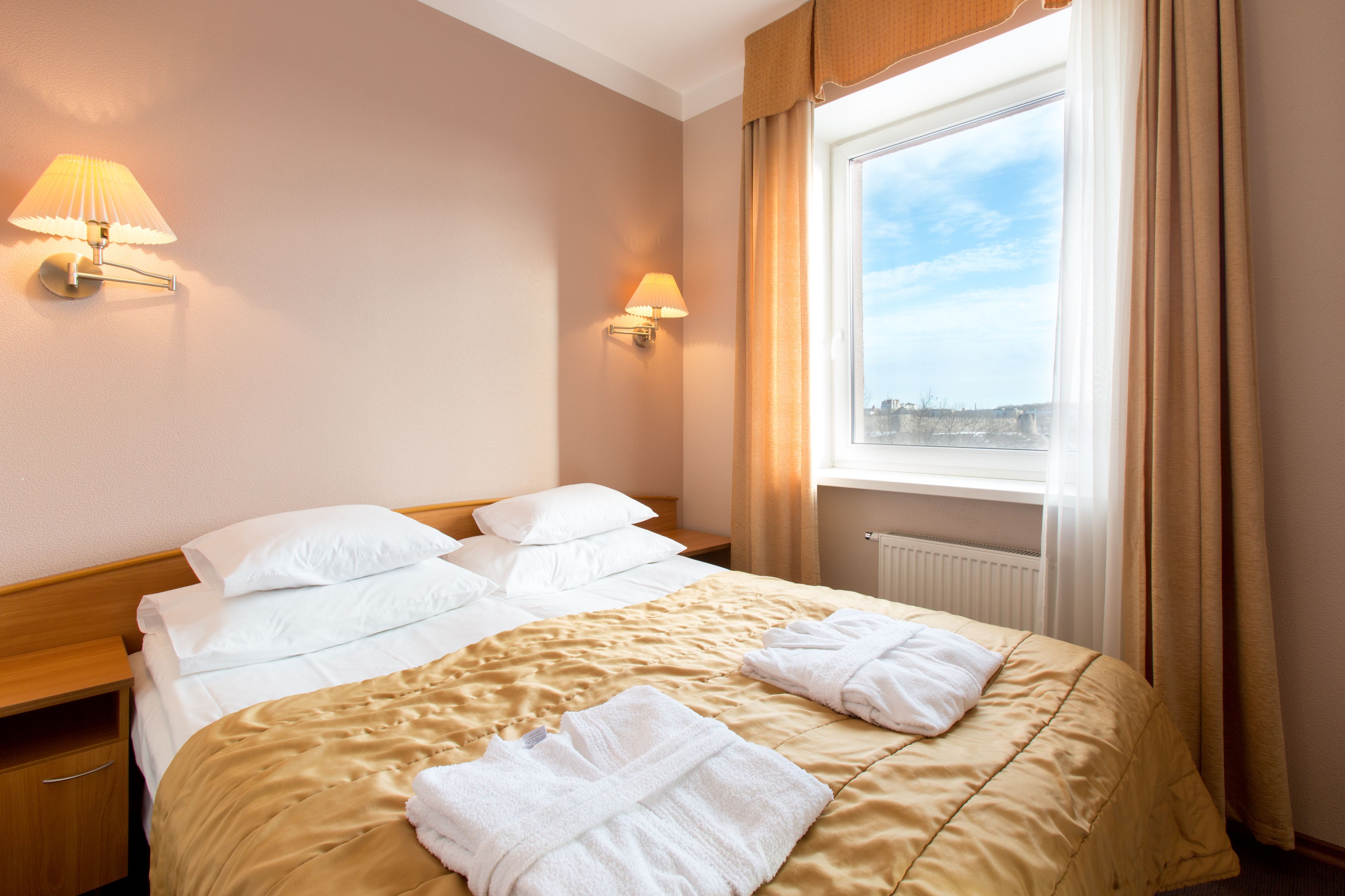 A hotel room with a bed, two bedside lamps, a window with curtains, and a view of the city.