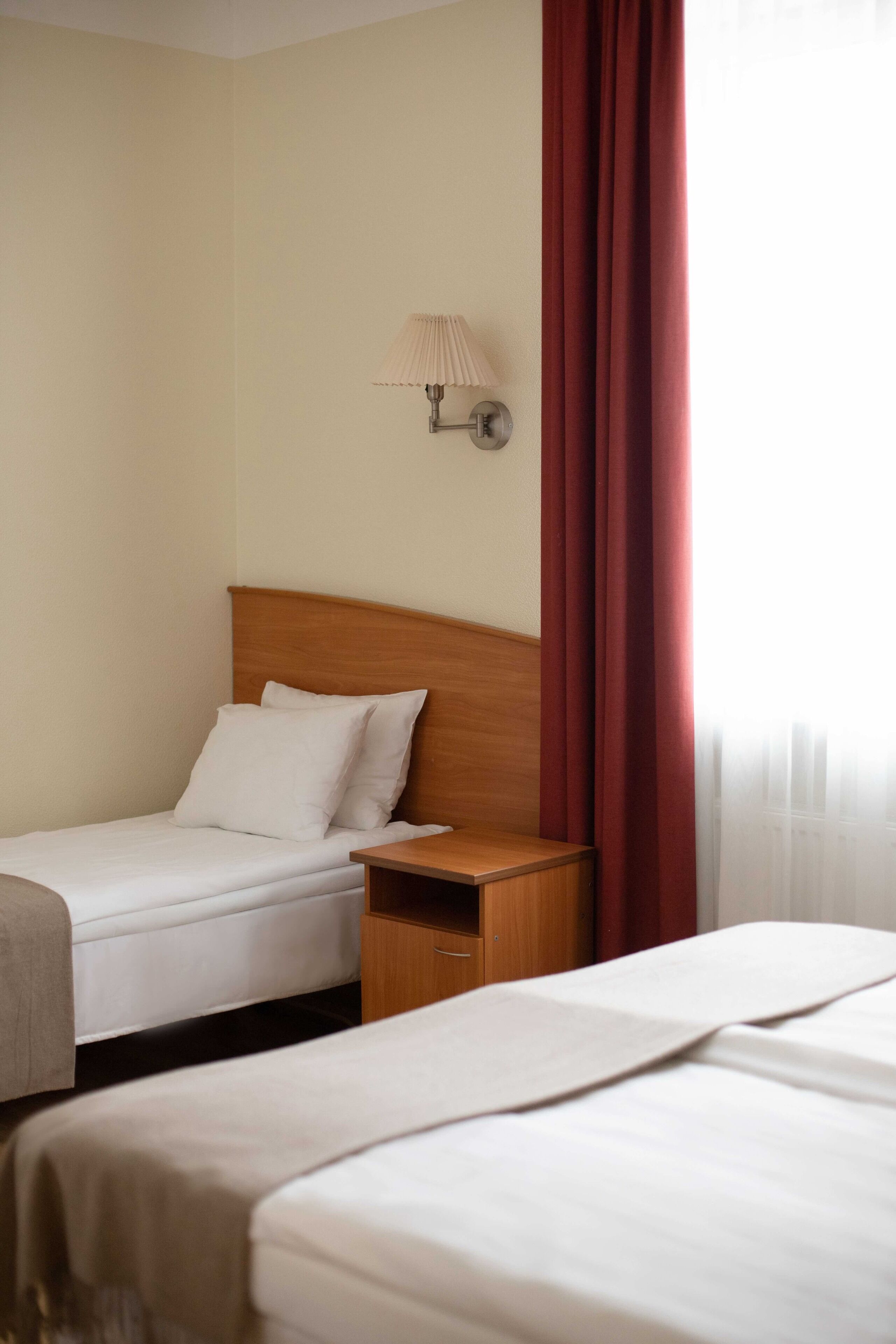 A hotel room with two beds, a wooden nightstand, a wall-mounted lamp, and a red curtain.