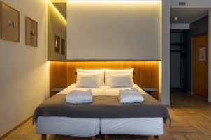 Una habitación de hotel con una cama grande, cabecera de madera y un espejo encima. Hay dos mesitas de noche y una pequeña mesita auxiliar.