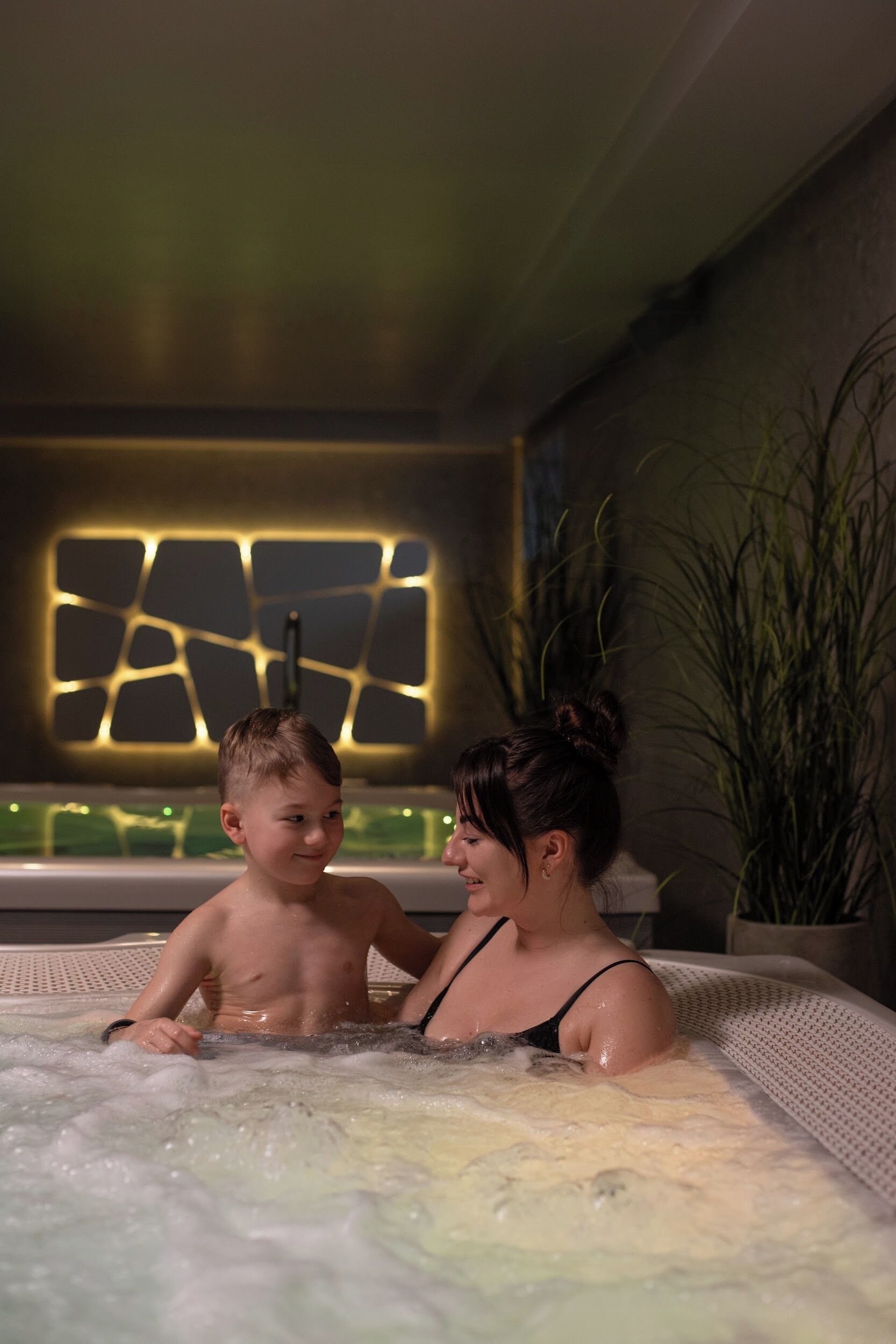 En kvinna och ett barn i ett bubbelbad, med en modern väggdesign och en växt i bakgrunden.
