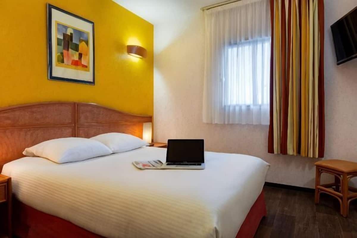 Habitación de hotel con cama, computadora portátil, mesita de noche, silla, un cuadro en la pared y ventana con cortinas.