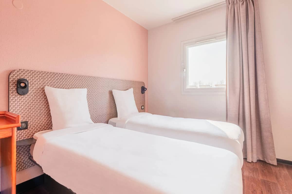 Habitación de hotel con dos camas individuales, una mesita de noche de madera, una ventana con cortinas y una pared con textura.